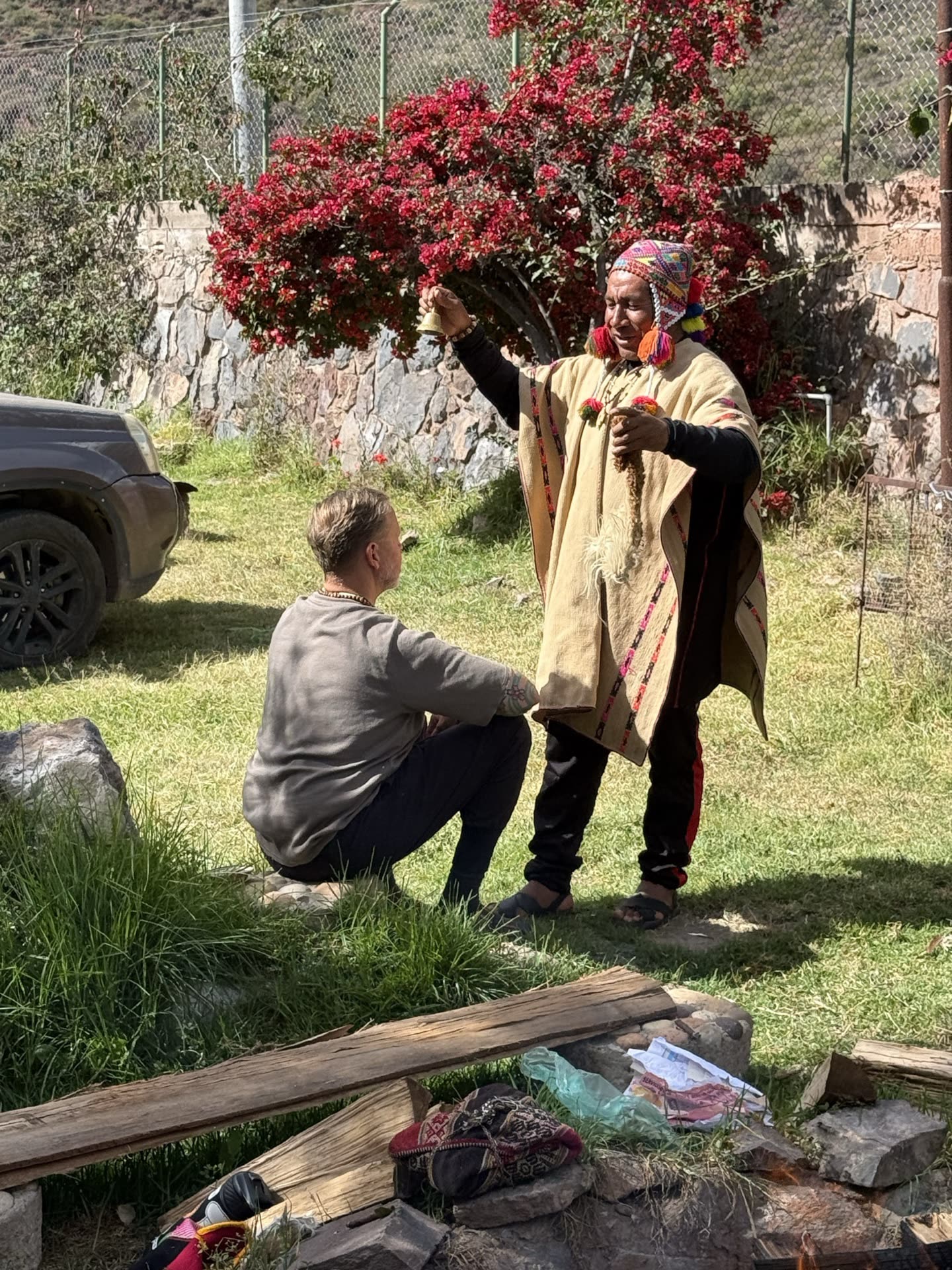 A shaman in traditional dress ringing a bell in blessing over a kneeling guest, bougainvillea behind.